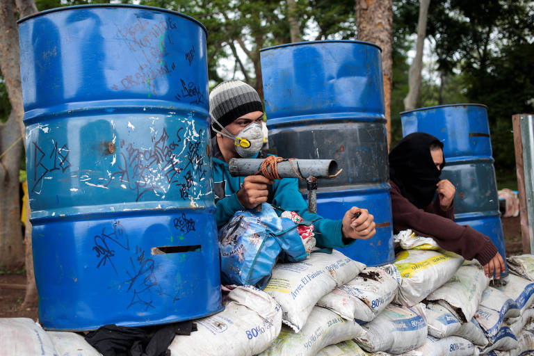 Dois manifestantes mascarados se posicionam em cima de sacos de areia brancos e entre tonéis de metal azuis. O primeiro deles da esquerda para a direita está com um morteiro caseiro na mão, enquanto o outro estica a mão entre os sacos de areia.