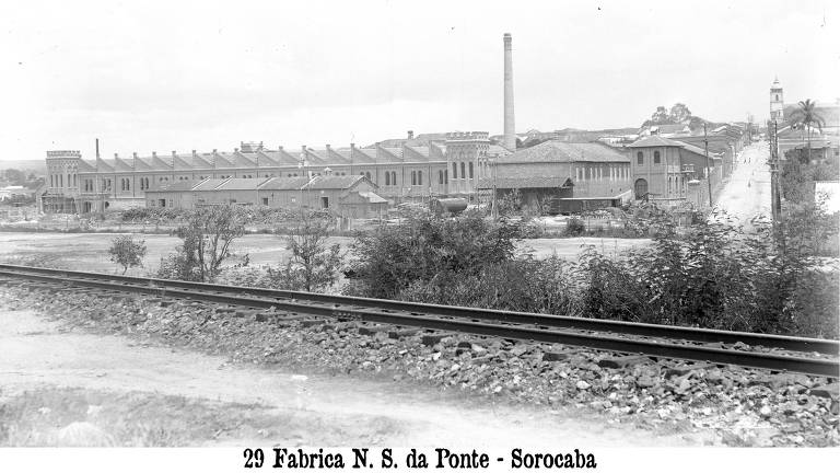 Foto em preto e branco do exterior da fábrica de tecelagem Nossa Senhora da Ponte. Na região, há apenas casas ou prédios baixos. Uma linha de trem passa ao lado da fábrica