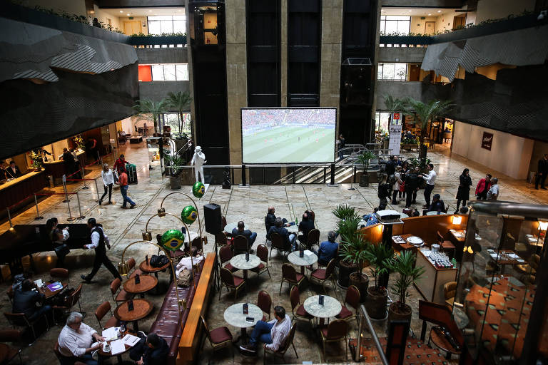 Imagem mostra vista aérea do lobby do hotel Maksoud Plaza. É um espaço grande, com várias mesas, vasos de plantas e escadas. Algumas pessoas andam por lá.