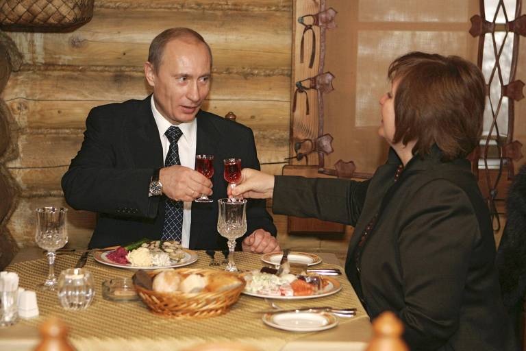 Com pães e comida sobre a mesa, Putin e sua mulher erguem um copo pequeno em um lugar de aparência simples, com paredes que lembram madeira