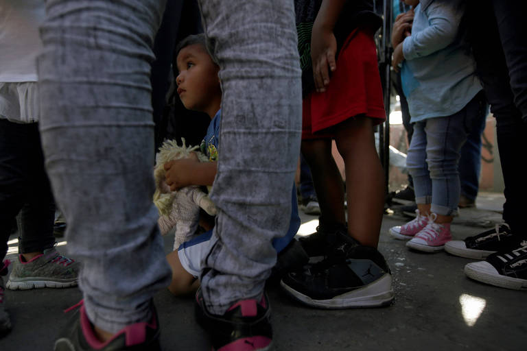 A criança aparece entre as pernas de uma mulher, que usa calça jeans preta. Ela segura um boneco de pano. Aparecem também as pernas de outras mulheres e de um homem.