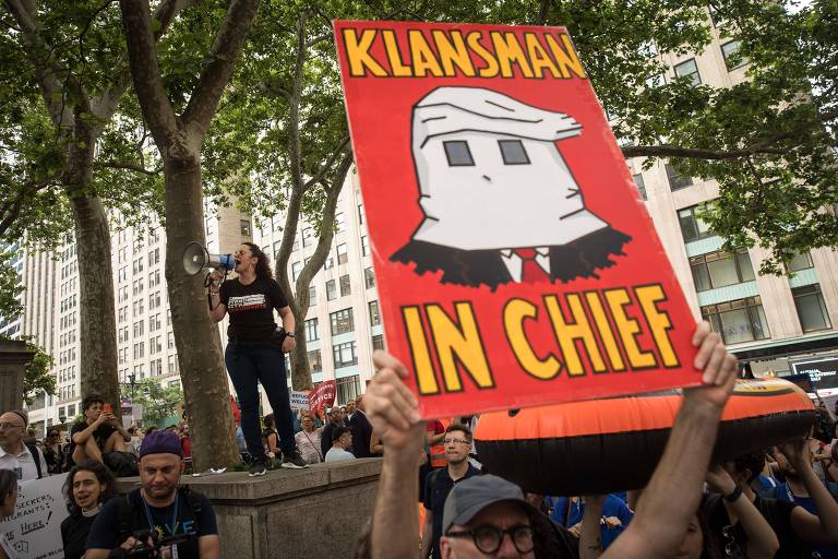 Homem de boné cinza carrega cartaz na horizontal em cor vermelha, com a expressão "Membro da Ku Klux Klan em chefe" em amarelo e uma caricatura de Trump no meio