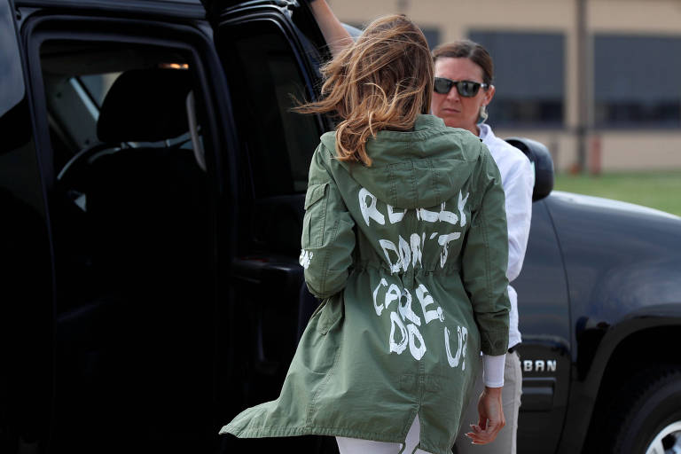 Melania aparece de costas, com a jaqueta verde, enquanto mulher abre porta de carro para que ela entre.