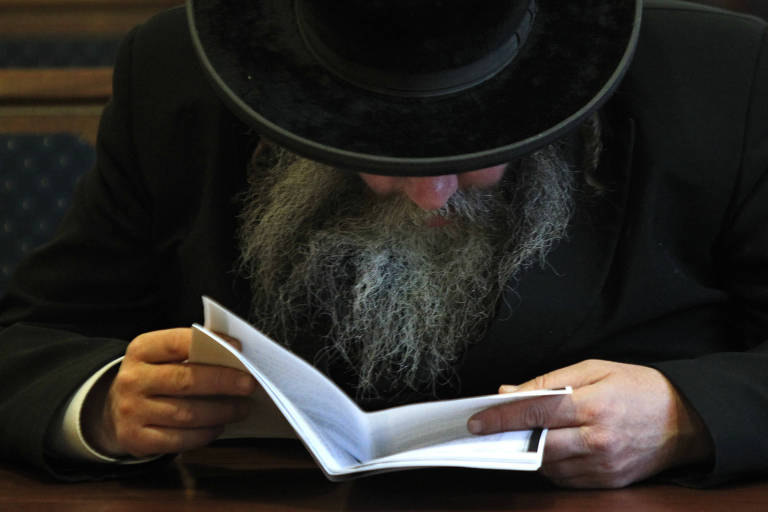 De roupas pretas, homem de barba longa está com a cabeça baixa lendo um pedaço de papel. O chapéu que usa encobre seu rosto, deixando aparecer apenas a ponta do nariz e a barba, um pouco grisalha. 