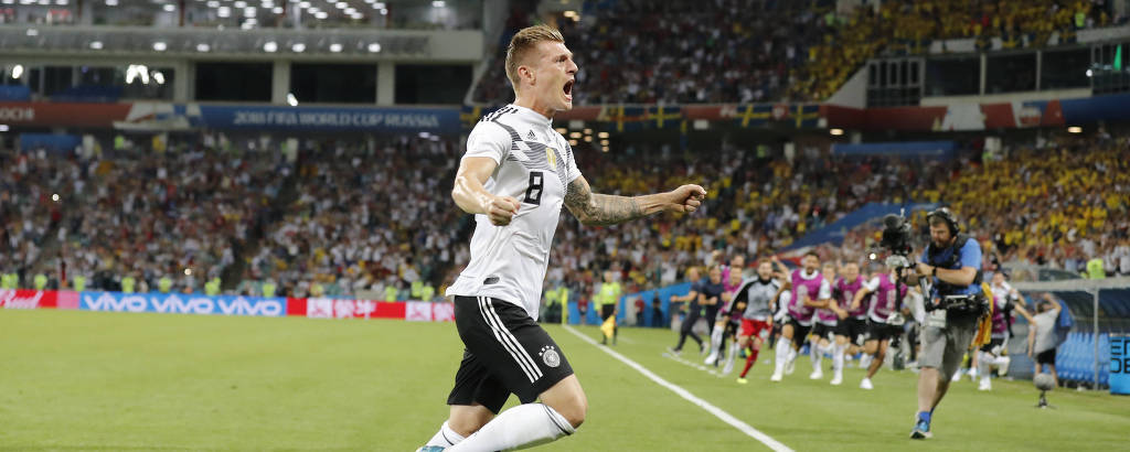 Toni Kroos, com uniforme branco da Alemanha, levanta os braços em comemoração ao gol marcado; ao fundo, companheiros de equipe no banco saem correndo ao encontro do jogador