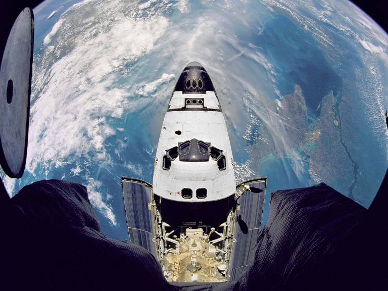 A parte frontal superior do ônibus espacial é vista de cima, com o planeta Terra ao fundo. A frente do ônibus espacial é pontuda e lembra a de um avião, com algumas janelas.