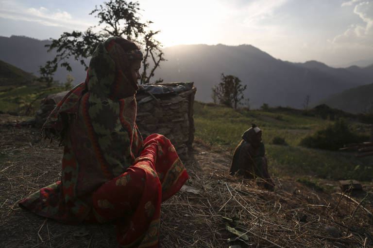 Mulher sentada em um gramado, na frente de uma cabana de pedras, com o sol e montanhas ao fundo