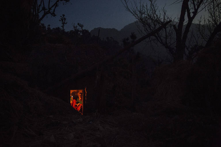 Imagem escura mostra montanhas e uma cabana com o interior iluminado, onde está uma mulher sentada