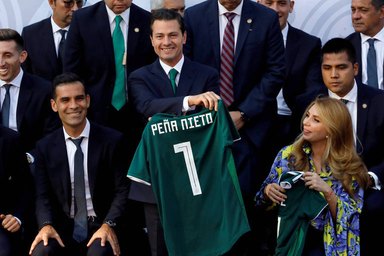 Sorrindo, Peña Nieto segura uma camisa verde com seu nome e o número 1. Ao lado direito dele, está Angelica Rivera, com vestido estampado e outra camisa verde na mão. Do lado direito aparece Rafael Márquez de terno, assim como os outros jogadores que estão atrás.