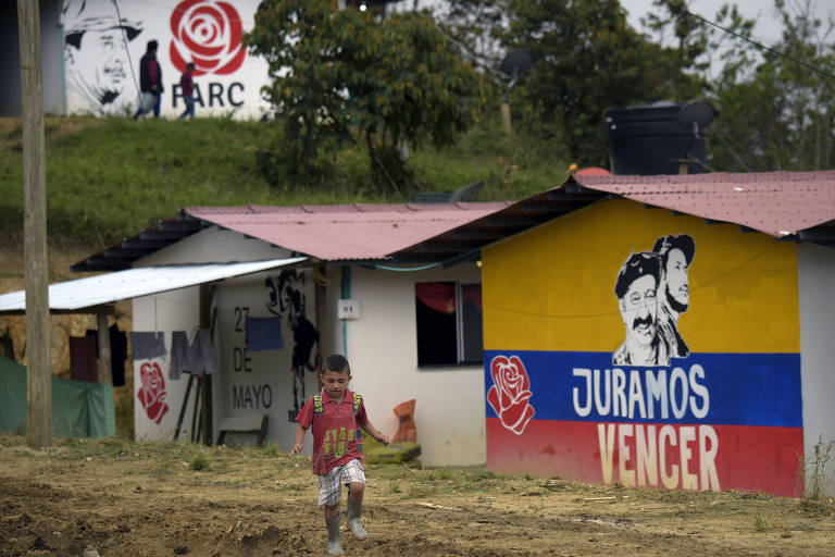Criança passa por área de terra e mato em um assentamento. À direita dele, duas casas, uma com um mural com a bandeira colombiana, o rosto de dois guerrilheiros, e a inscrição "Juramos Vencer". Acima das casas, um muro sobre um barranco traz a rosa e a inscrição FARC, do novo partido político.