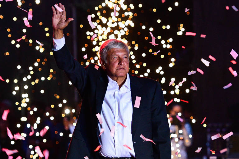 De blazer preto e camisa branca, López Obrador levanta a mão esquerda para se despedir de seus seguidores. Ao fundo, caem papéis picados brancos, vermelhos e cor de rosa e aparecem fogos de artifício.