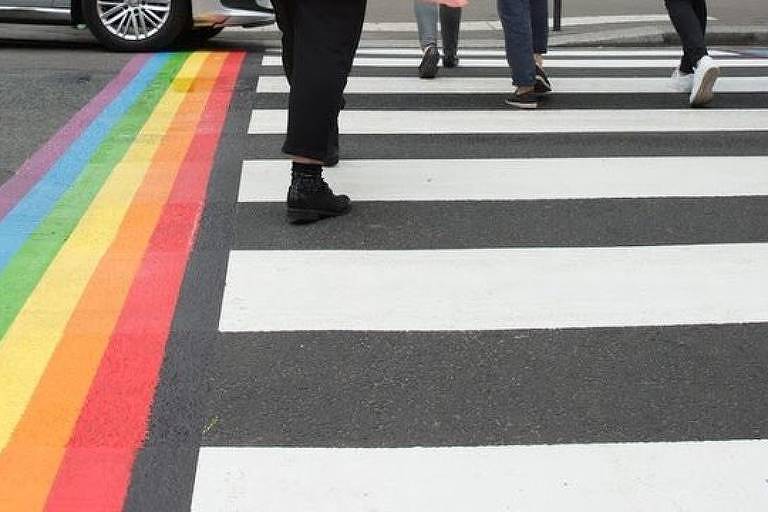 Pessoas atravessam uma faixa de pedestres. No lado esquerdo dela há uma faixa do arco-íris perpendicular.