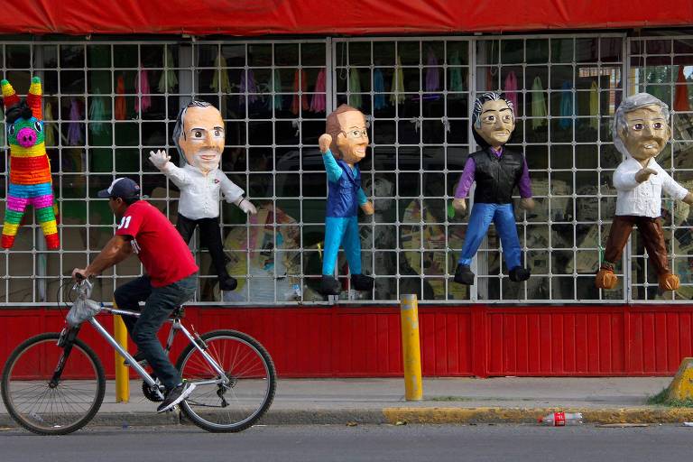 Os bonecos aparecem pendurados em uma grade de loja em formato de quadrado. À esquerda deles aparece uma piñata colorida de um burro. Um homem passa de bicicleta pela frente da loja.