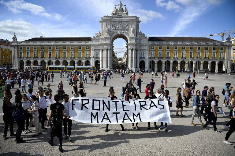 Grupo de pessoas carregam manta branca com a inscrição em preto Fronteiras Matam em uma praça em frente a um prédio histórico de Lisboa com um pórtico no meio.