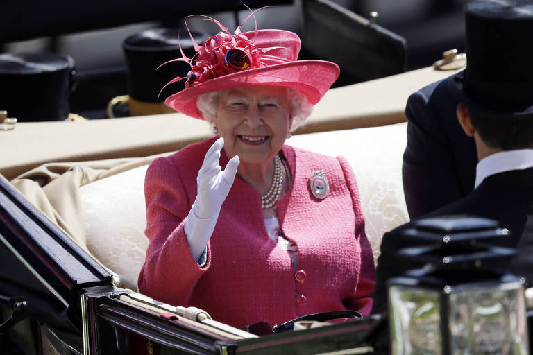 Elizabeth 2ª sorri de dentro de uma carruagem aberta. Ela acena para o público com a mão esquerda, calçada com uma luva branca, e usa um terninho rosa com um chapéu da mesma cor.