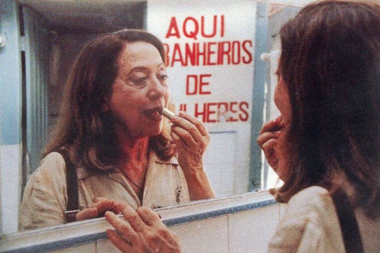 Em cena do filme, Fernanda Montenegro está em um banheiro público, diante de um espelho, passando batom na boca 