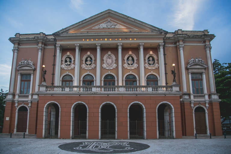 A imagem mostra a fachada de um teatro com uma arquitetura clássica. A estrutura é de cor laranja e possui colunas brancas， janelas com arcos e detalhes decorativos. Acima das janelas， há retratos emoldurados. O céu ao fundo está claro， indicando um horário do dia próximo ao entardecer.