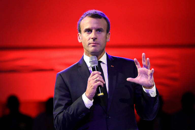 Retratado da cintura para cima, Macron aparece gesticulando com a mão esquerda aberta, enquanto segura o microfone com a direita. O fundo do cenário de onde ele fala é vermelho.