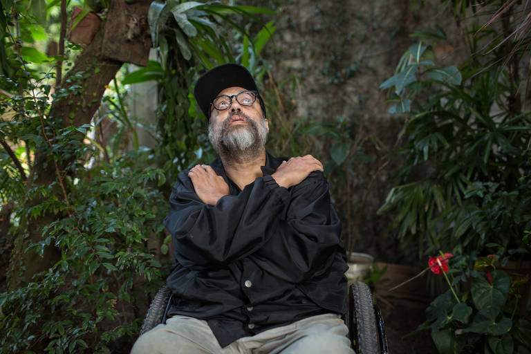 Marcelo Yuka sentado em cadeira de rodas cruza os braços na frente de árvores