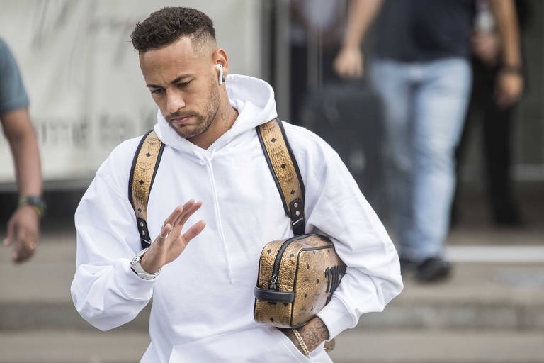 Neymar, com expressão abatida, levanta uma mão como quem diz "agora não"; ele veste um moletom branco, está com uma necessaire dourada e bolsa dourada escrito "nrj"