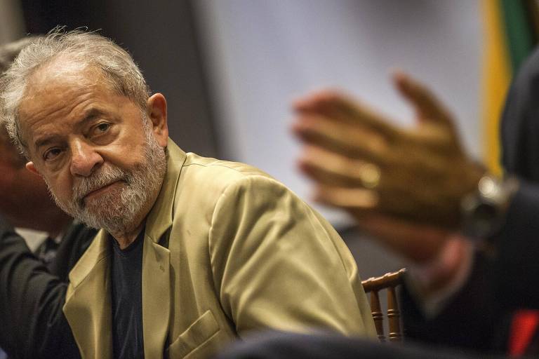 Lula se inclina ao fundo, em primeiro plano, mãos batem palmas