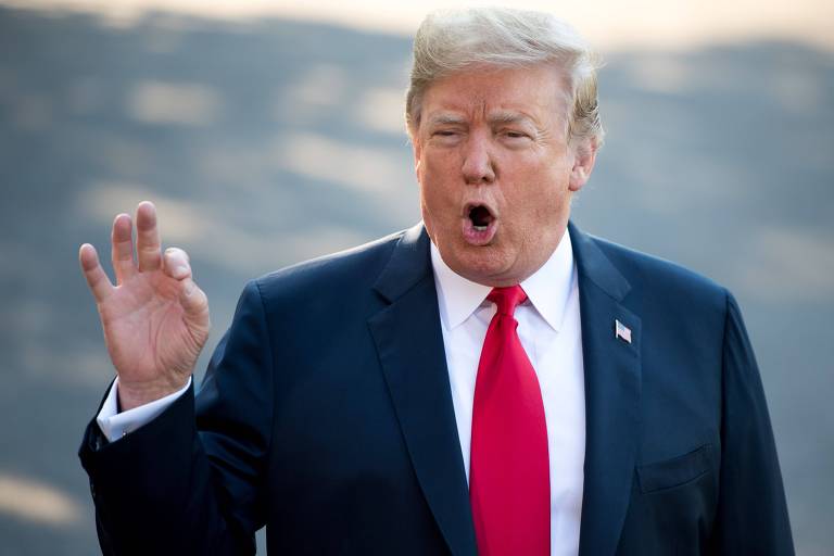 De terno preto, camisa branca e gravata vermelha, Trump gesticula com a mão esquerda enquanto fala. Ele aparece da cintura para cima.