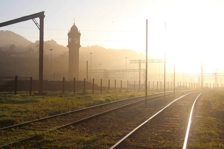 Uma linha de trem no meio do campo aparece em primeiro plano. Ao fundo, contra a luz do sol, a torre da estação de Paranapiacaba. Atrás dela, parte da serra