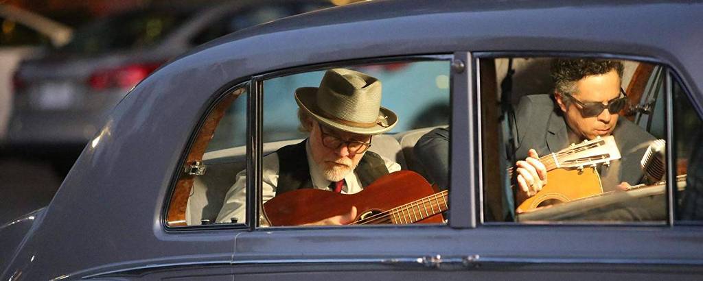 Dois músicos, um velhinho, de chapéu, e um mais novo, de óculos escuros, tocam dentro de um carro antigo