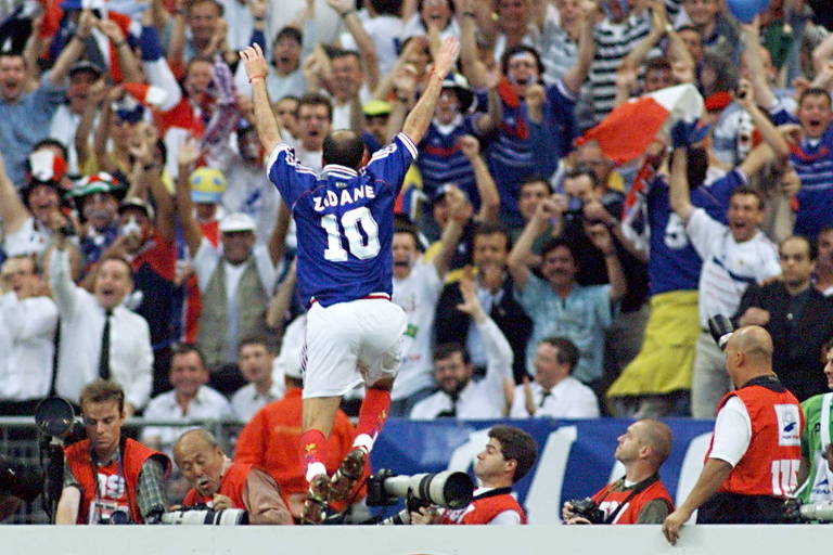 Usando a camisa 10 da França e com os braços erguidos, Zidane salta uma placa publicitária para ir na direção da torcida a fim de comemorar o primeiro de seus dois gols na final da Copa de 1998, contra o Brasil, no Stade de France   