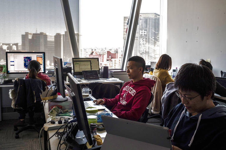quatro pessoas trabalham em seus computadores, ao fundo, ampla janela de vidro mostra os prédios da cidade