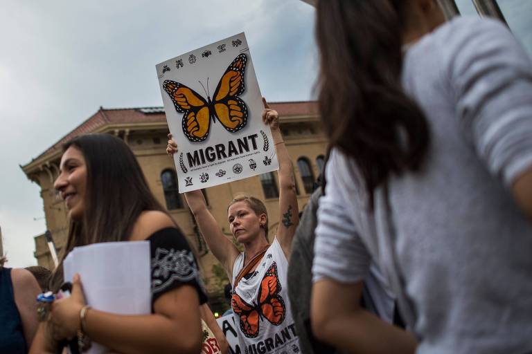 Mulher carrega cartaz com borboleta e a palavra imigrante. Ela usa uma camiseta com a mesma estampa.