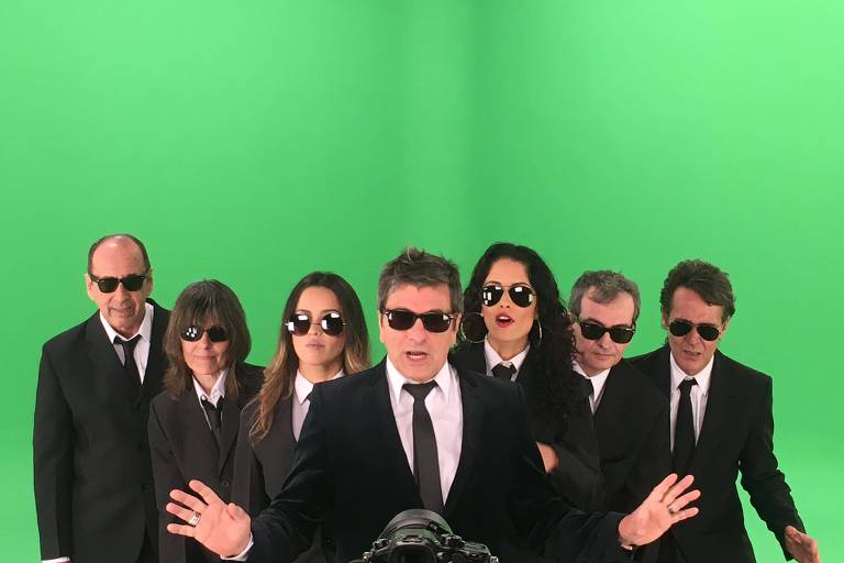 Músicos da banda Blitz estão vestidos com roupas pretas e óculos escuros em foto de fundo verde