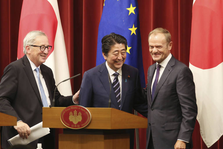 Os três, de terno, aparecem atrás de um púlpito com um microfone. Atrás deles estão duas bandeiras japonesas e uma da União Europeia no meio, cercadas por uma cortina vermelha.