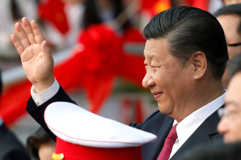 Xi Jinping levanta a mão e sorri. Ele aparece de perfil entre assessores e militares (o quepe branco e vermelho de um deles aparece na imagem).