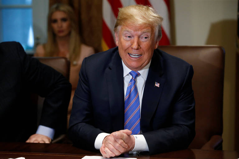 De terno preto, camisa branca e gravata azul com listras vermelhas, Trump está sentado a uma mesa, de boca aberta como se estivesse falando, e com as mãos unidas.
