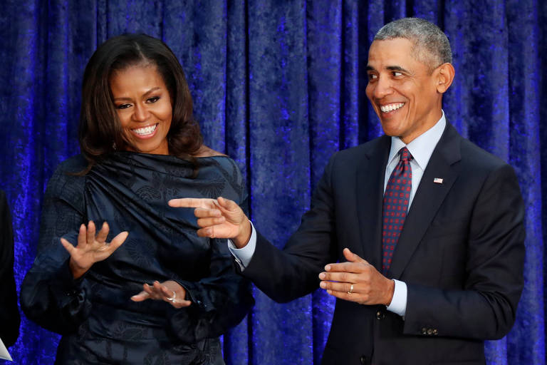 Obama, de terno preto, camisa cinza e gravata vinho e azul, e Michelle, de vestido cinza escuro, riem. O ex-presidente, que está à direita, aponta com o dedo indicador esquerdo para um local que não está na imagem. Michelle, que está à esquerda, faz um sinal com a mão esquerda. Os dois estão em um local com cortinas azuis escuras no fundo.