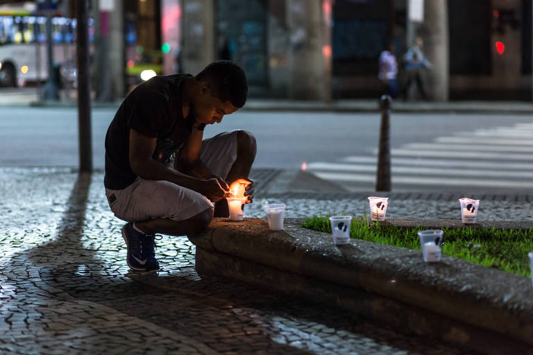 a foto mostra um jovem que se ajoelha diante velas posicionadas numa rua no centro do rio de janeiro, à noite