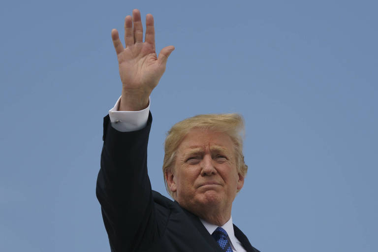 De terno preto, camisa branca e gravata azul, Trump levanta a mão para o alto. Ele aparece de lado, do ombro para cima.