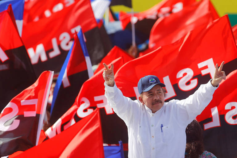 De camisa branca e boné azul, Daniel Ortega levanta os braços e faz o "v" da vitória com nas duas mãos. Ao fundo, aliados agiram bandeiras vermelhas e pretas da frente sandinista.