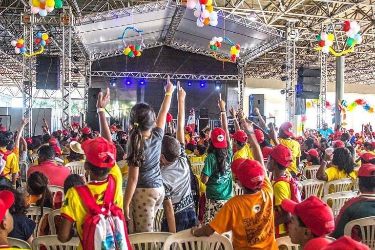 Crianças que vivem em assentamentos e acampamentos do MST durante encontro "sem terrinha", em Brasília; As crianças erguem os dedos para a cima em um evento em um galpão