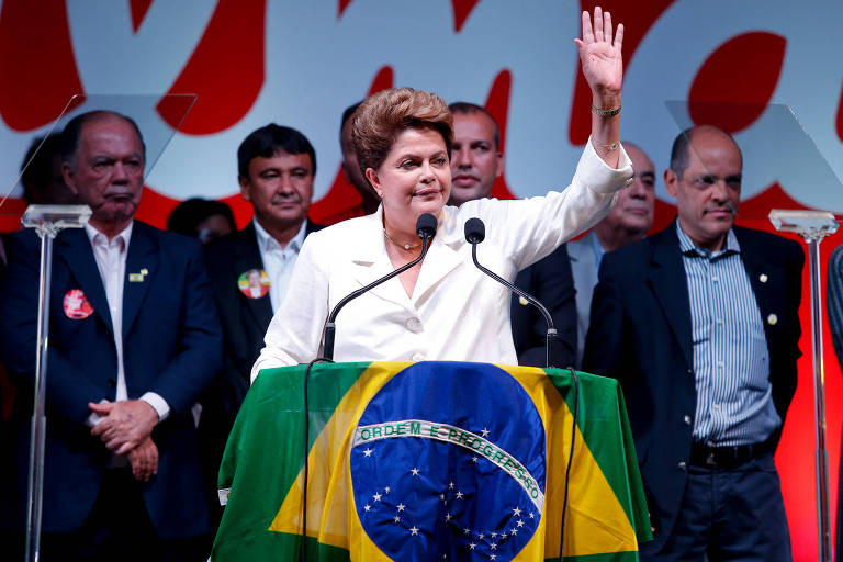 Dilma aparece acenando. Ela está no palanque, discursando. Veste paletó branco e está cercada de políticos que preenchem o fundo da imagem. O pódio no qual discursa está coberto por uma bandeira do Brasil.
