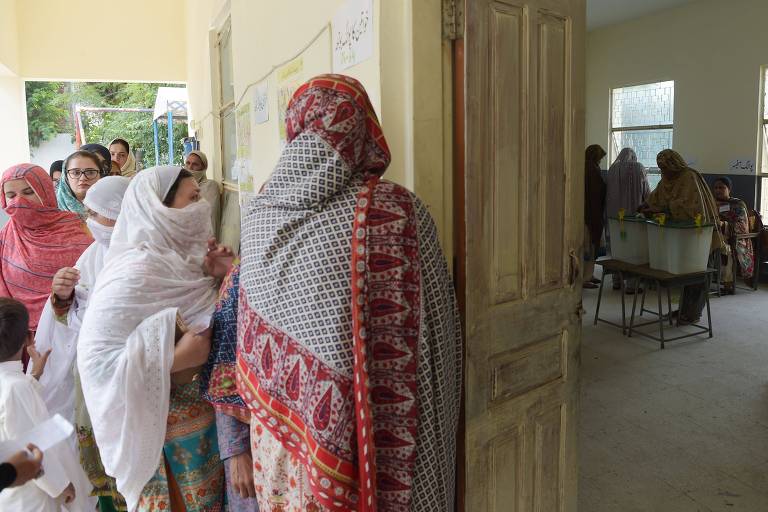 À esquerda há um grupo de seis mulheres, todas usando túnicas e véus islâmicos de diferentes cores, fazendo fila na porta de uma sala. Dentro da sala, que se vê do lado esquerdo, duas mulheres votam ao longe.