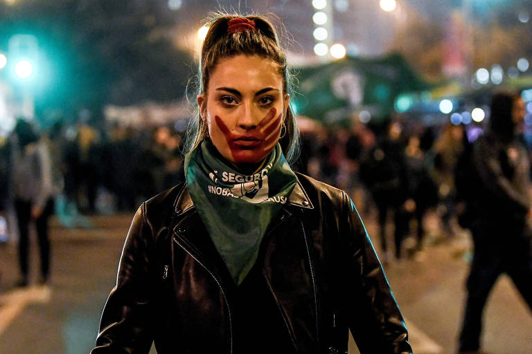 De roupa preta, mulher usa lenço verde no pescoço e pintou seu rosto com uma mão vermelha, simulando um tapa. Ao fundo, outras pessoas protestam.