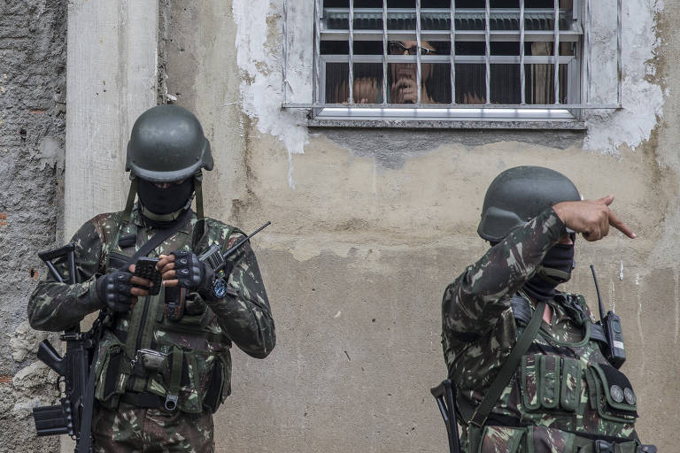 Soldados com uniforme camuflado e capacete escuro estão em frente à parede pintada em tom de marrom claro com reboco e janela com grade onde está uma pessoa
