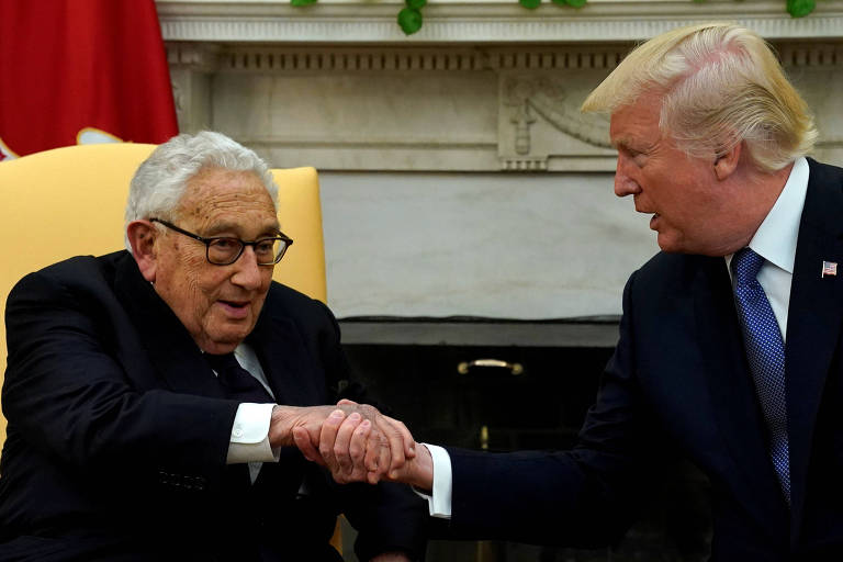 Trump usa terno preto e está à direita da imagem, estendendo sua mão direita e apertando a mão de Kissinger, que também usa terno. Os dois estão sentados em poltronas em frente a uma lareira.