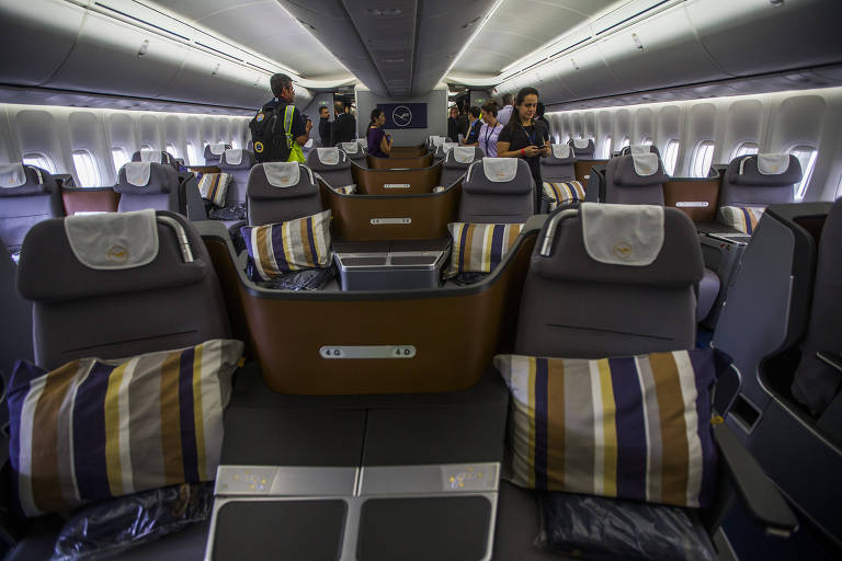 Imagem mostra assentos acolchoados com divisórias em um avião. Passageiros circulam pelo corredor.