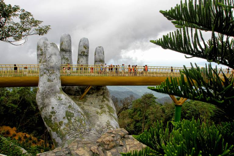 Turistas andam por ponte no Vietnan que tem duas mãos de concreto segurando a obra
