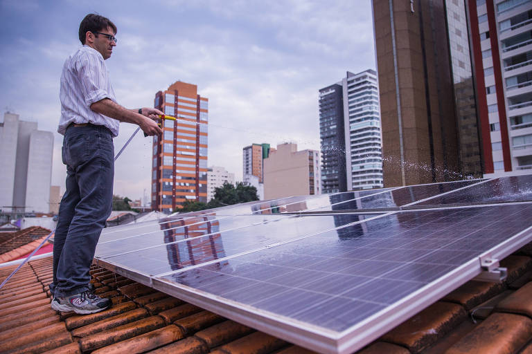 Homem está em pé no telhado em frente a um painel solar. Em volta, há vários prédios.