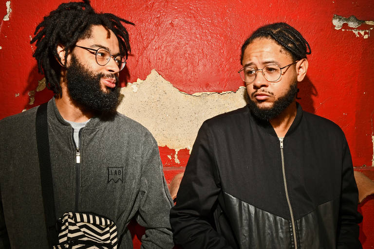 A imagem mostra dois homens em pé contra uma parede de cor vermelha desgastada. O homem à esquerda tem cabelo crespo e barba， usa óculos e uma jaqueta cinza. O homem à direita também tem cabelo crespo， usa óculos e uma jaqueta preta. Ambos têm expressões faciais que sugerem uma conversa ou interação.