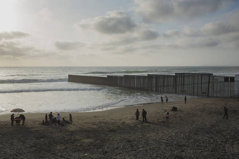 um praia, pessoas na praia e, partindo da areia e avançando ao mar, um muro